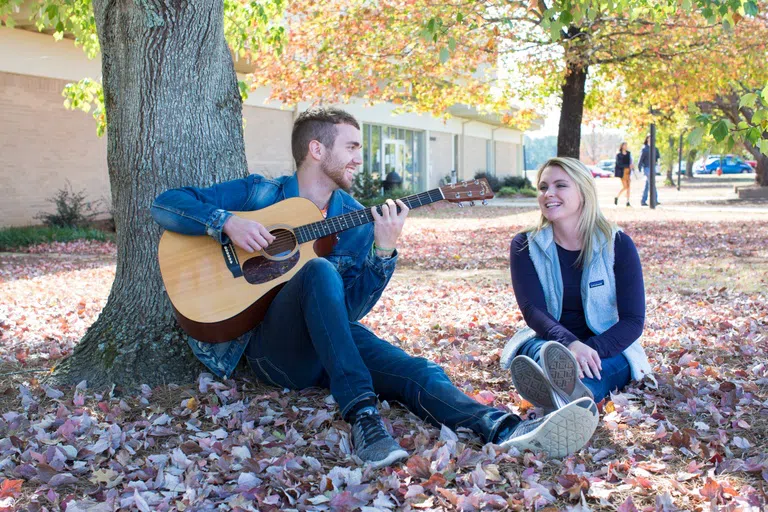 Two students sit in the grass outside the Floyd Library. One is playing guitar while the other listens