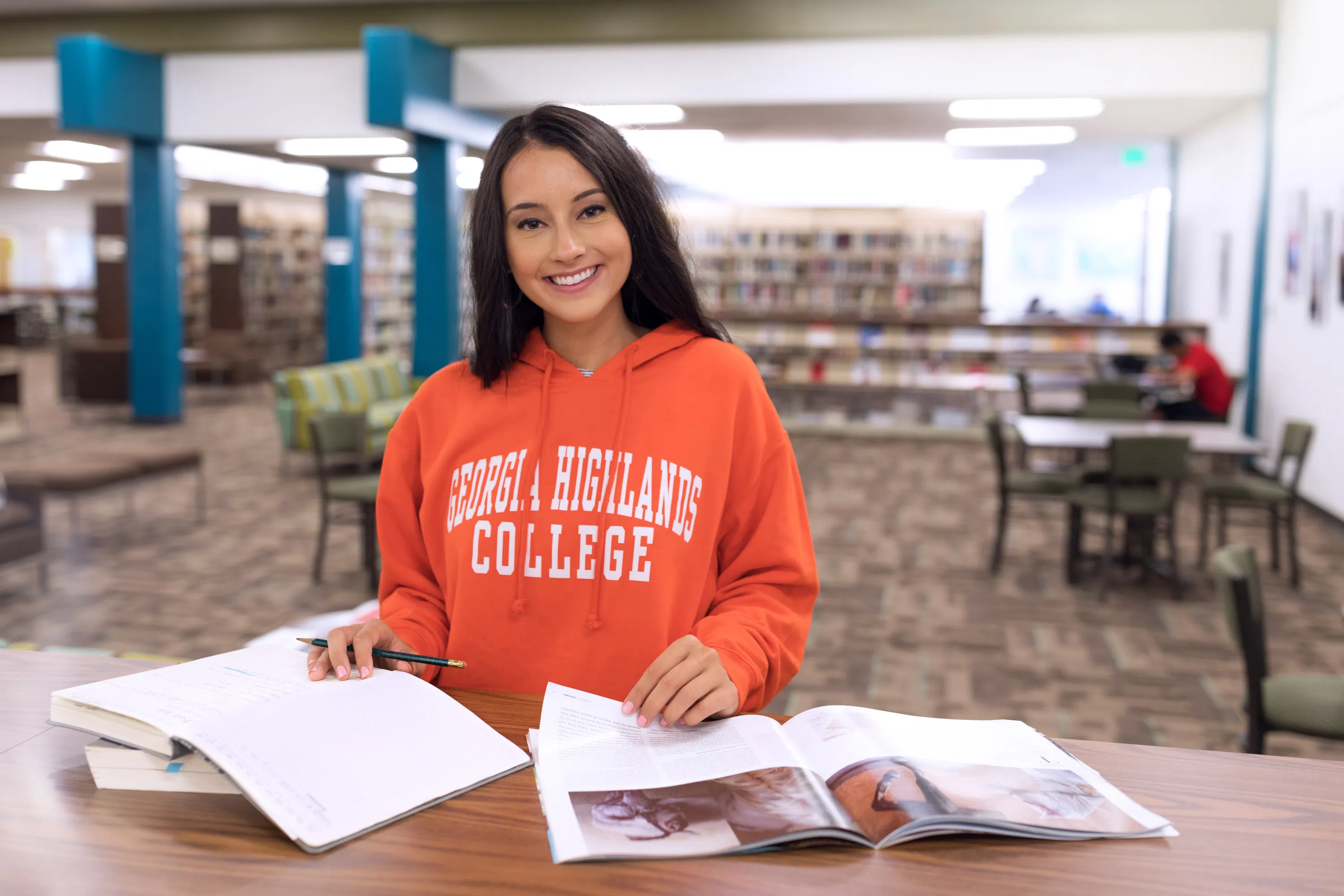 Student wearing an orange Georgia Highlands sweatshirt looks up from a book and smiles at the camera