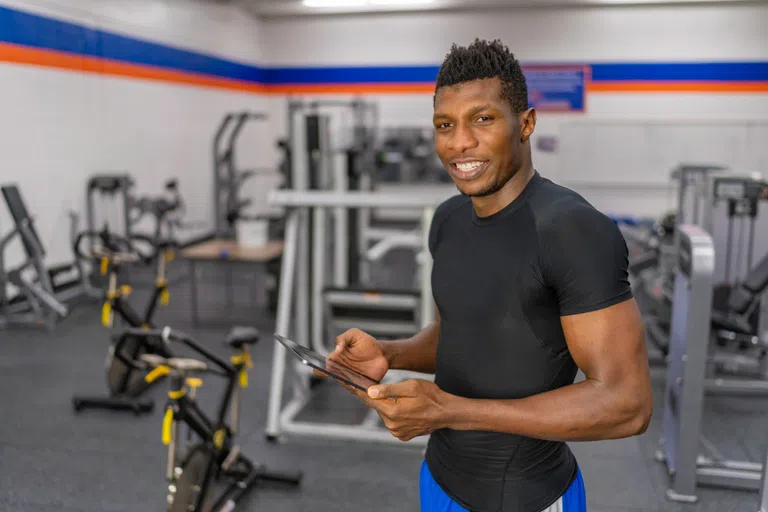 Trainer stands in the Physical Education Fitness Center with an iPad. Exercise equipment placed throughout the room in the background