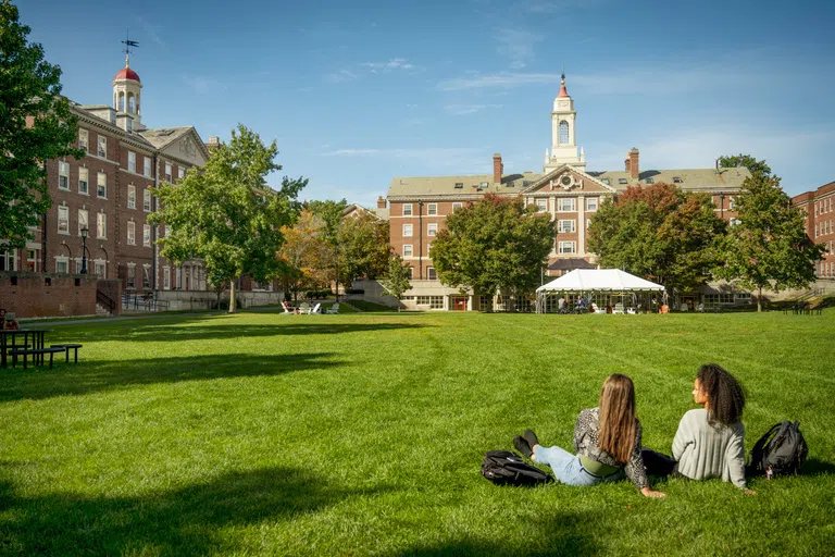 Two students sit on a grassy lawn, spired building and trees in the background