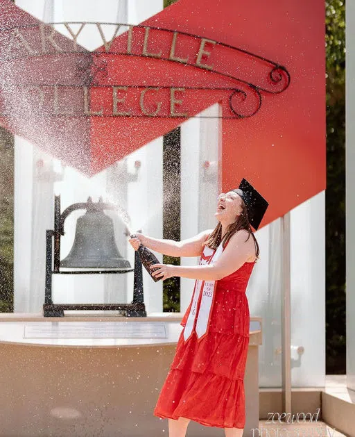 A student pops champagne in front of the Maryville Bell and the "Maryville College" sign with the Big Red "M"