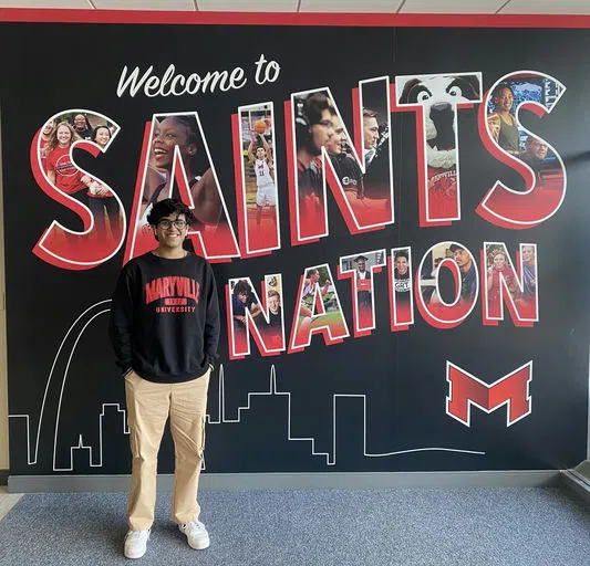 A student in a black Maryville sweatshirt stands in front of the "Welcome to Saints Nation" wall.