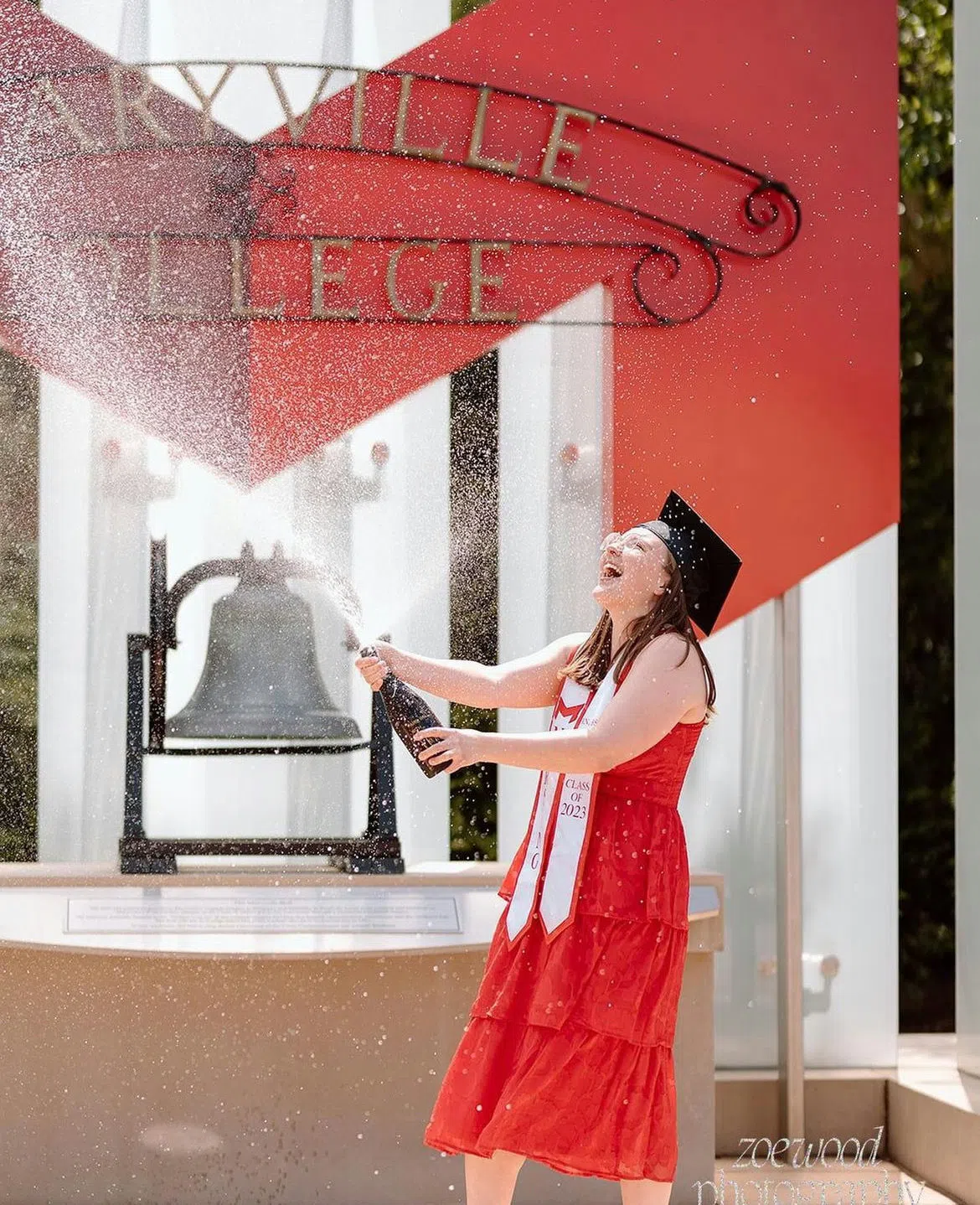 A student pops champagne in front of the Maryville Bell and the "Maryville College" sign with the Big Red "M"