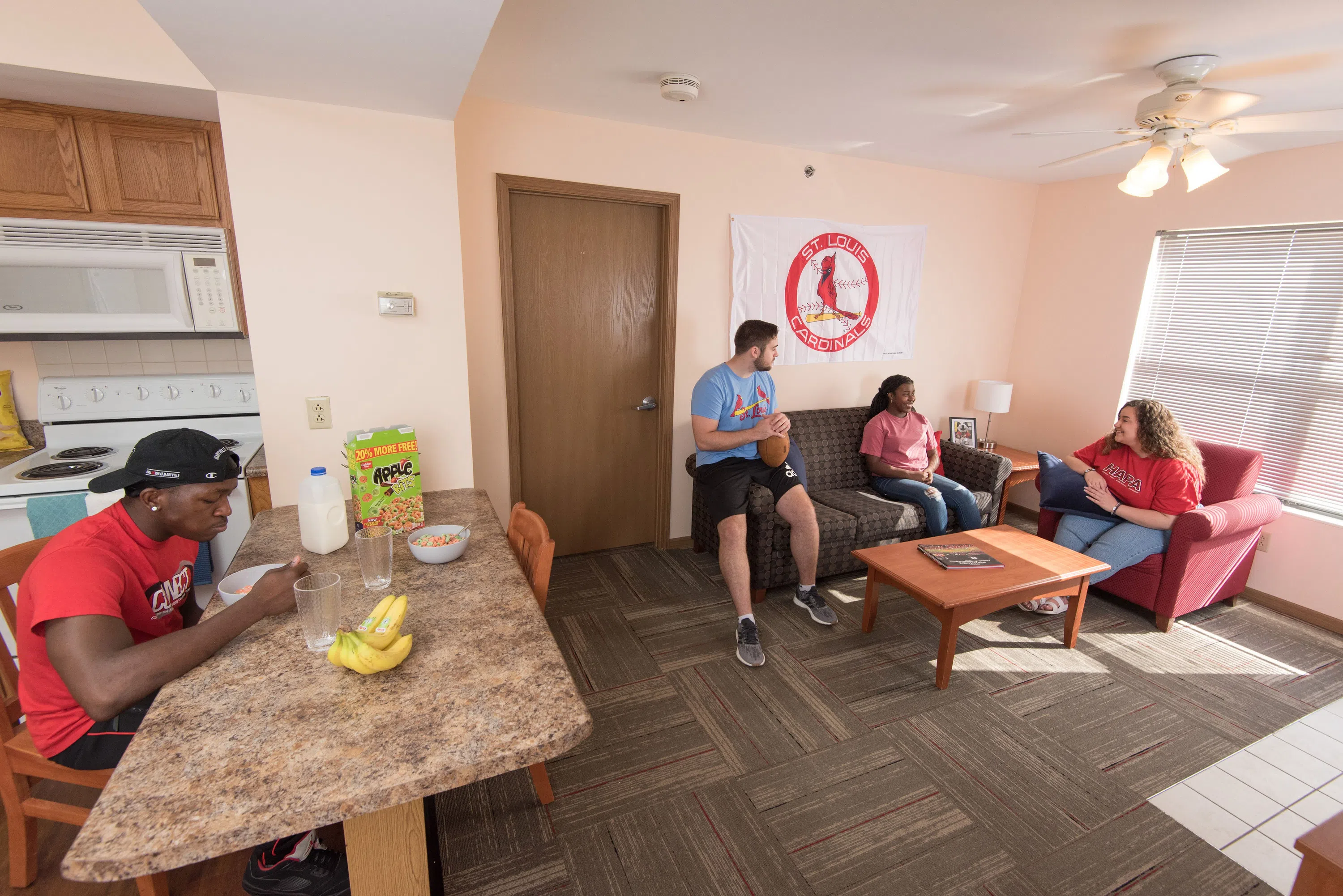Students hang out together in an on-campus apartment. Some sit on a couch, watching TV and talking. Another student sits at the kitchen counter to eat.