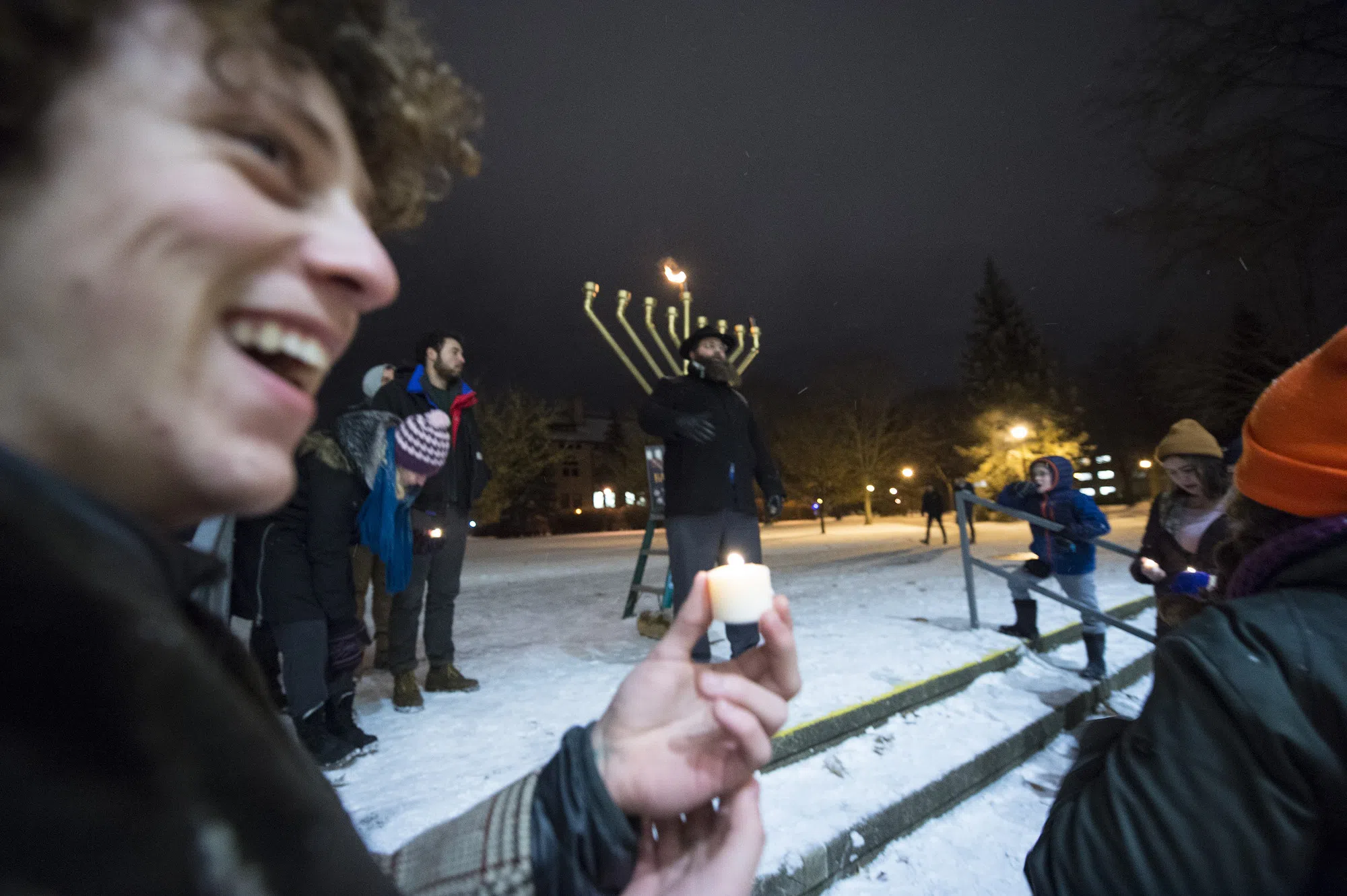 A student smiles with a small candle as the large menorah for Hanukkah is lit in the background.