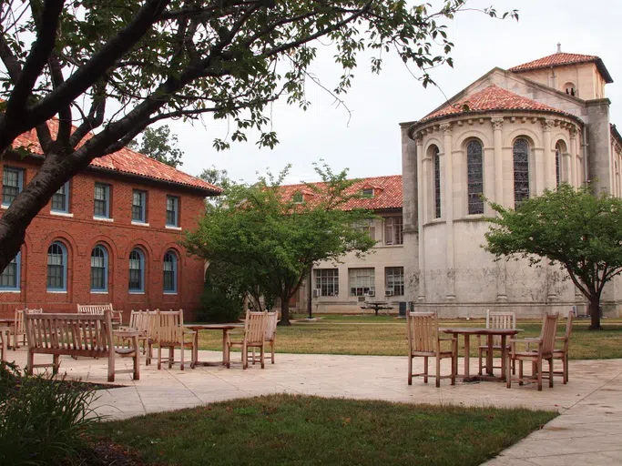 A large red brick building encloses a peaceful courtyard with tables and chairs. 