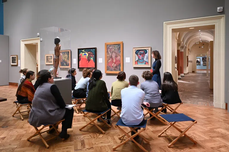 A student presents on a piece of art work in front of an audience of 12 people sitting in blue fold-up chairs.