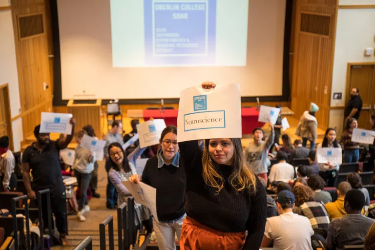 Students hold up signs with majors in a large lecture hall. 