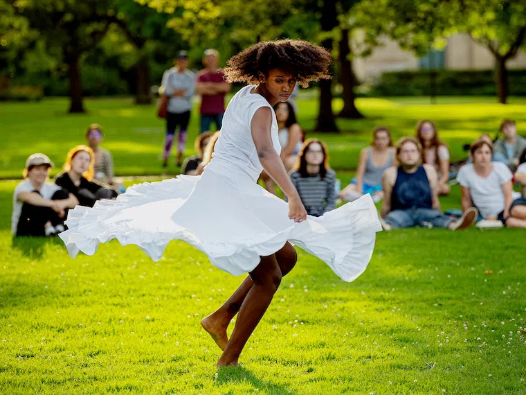 A student dances in a white dress in front of a small crowd of sitting students. 