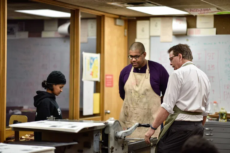 A professor and two students wearing aprons gather around the letter press. 