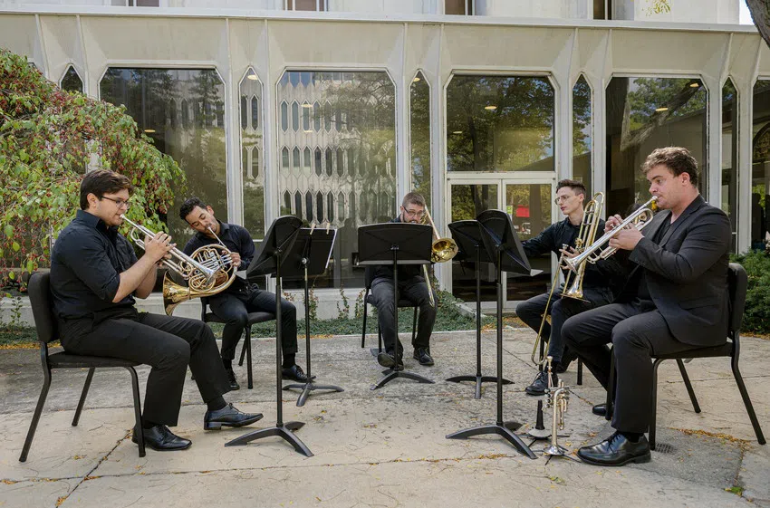 Five students in a horn ensemble of trumpet, french horn, and trombone, perform a quintet together outside a building with long hexagonal windows.