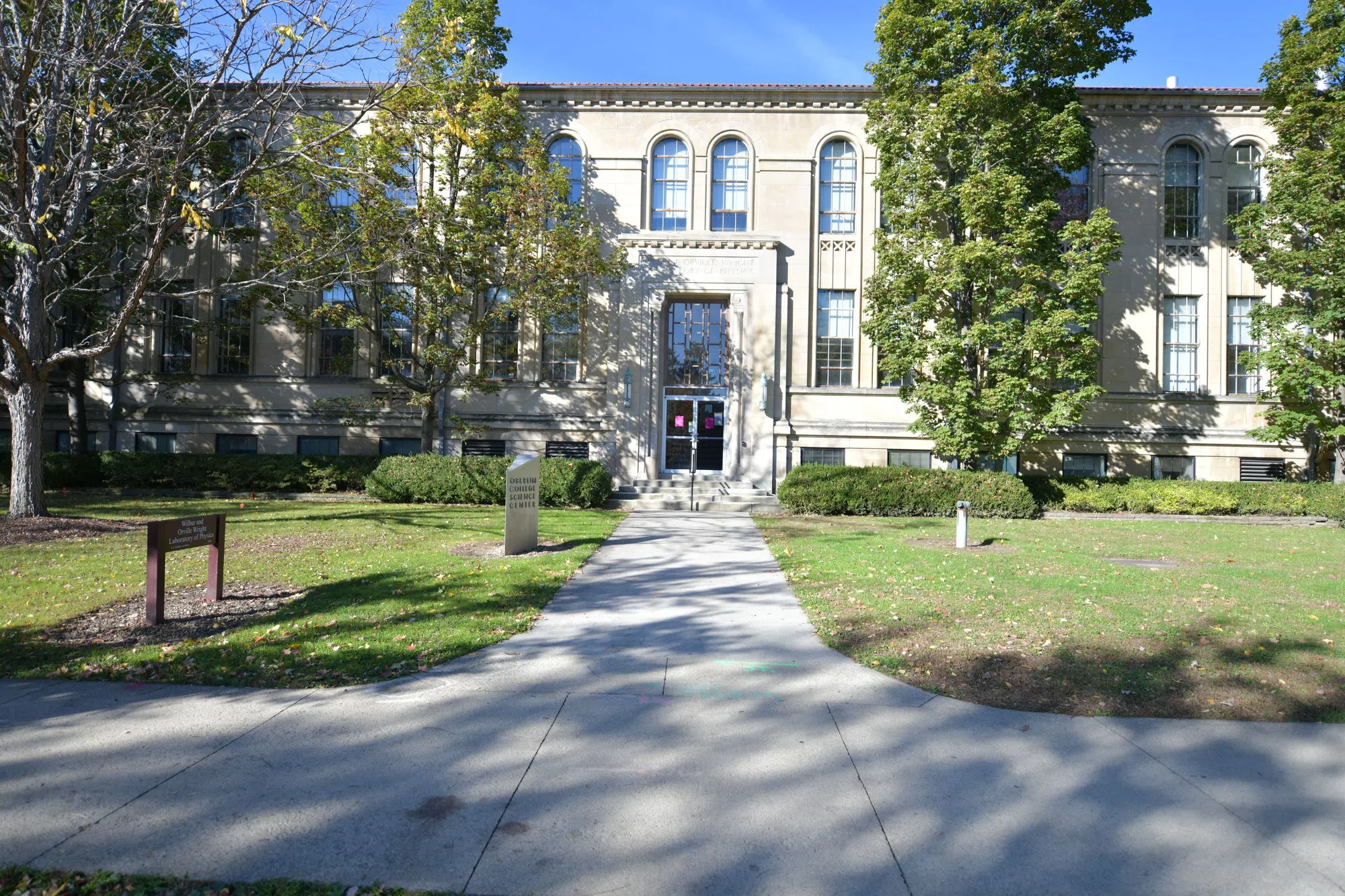 Pictured is a sandstone building from the early 1900s, known as the Wright Physics Lab building. Greenery surrounds the building. 