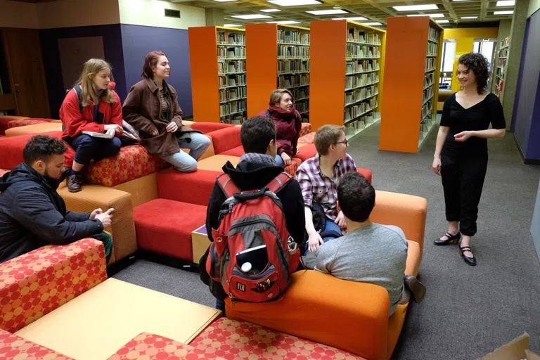 A group of students with backpacks sit on orange chairs facing a standing speaker.