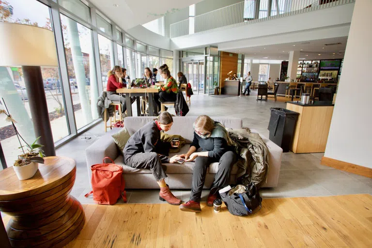 Two students sit on a couch. A cluster of students sit in the back at a table discussing something.