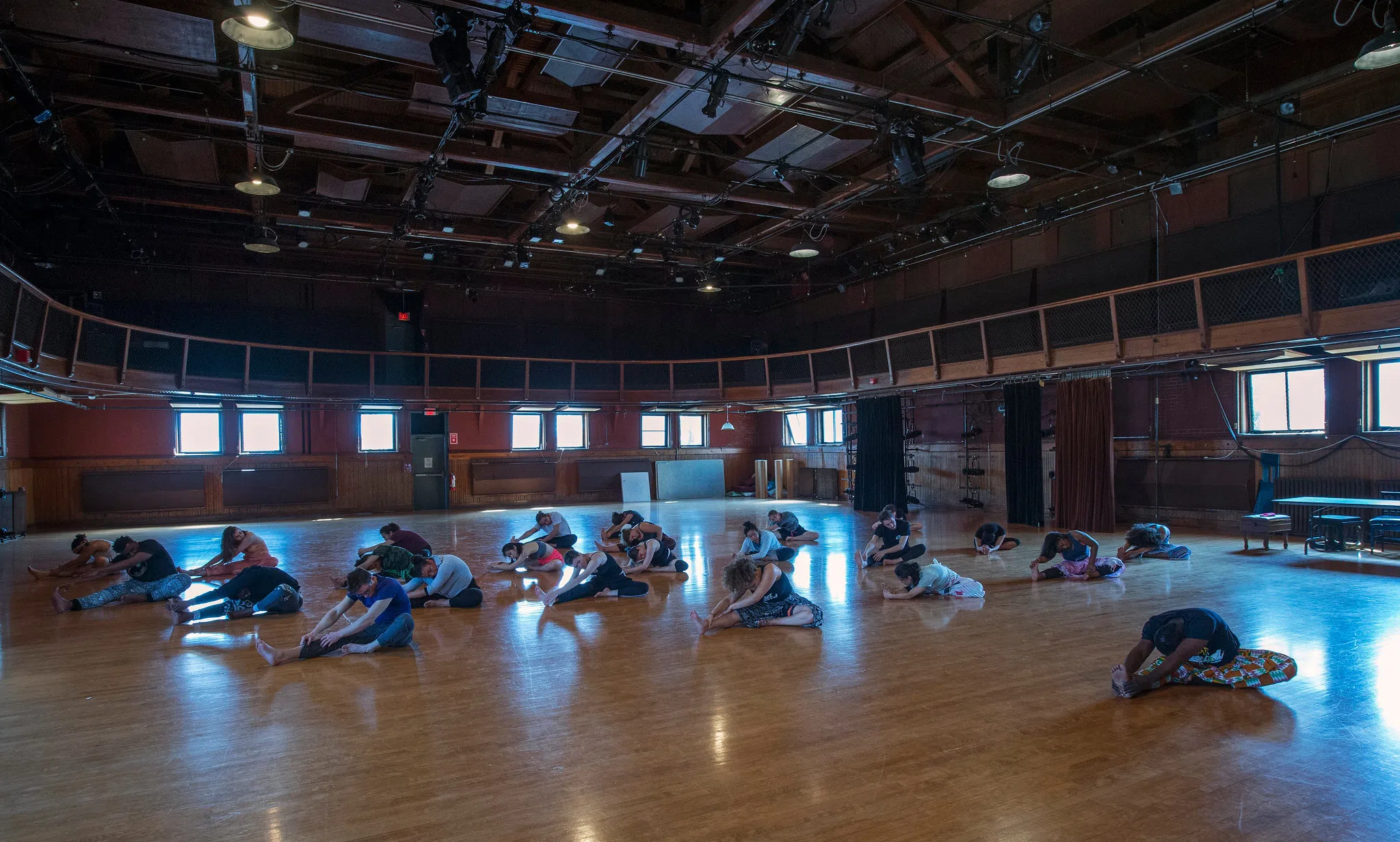 About 15 students and a dance professor stretch their legs in a wide dance studio. 