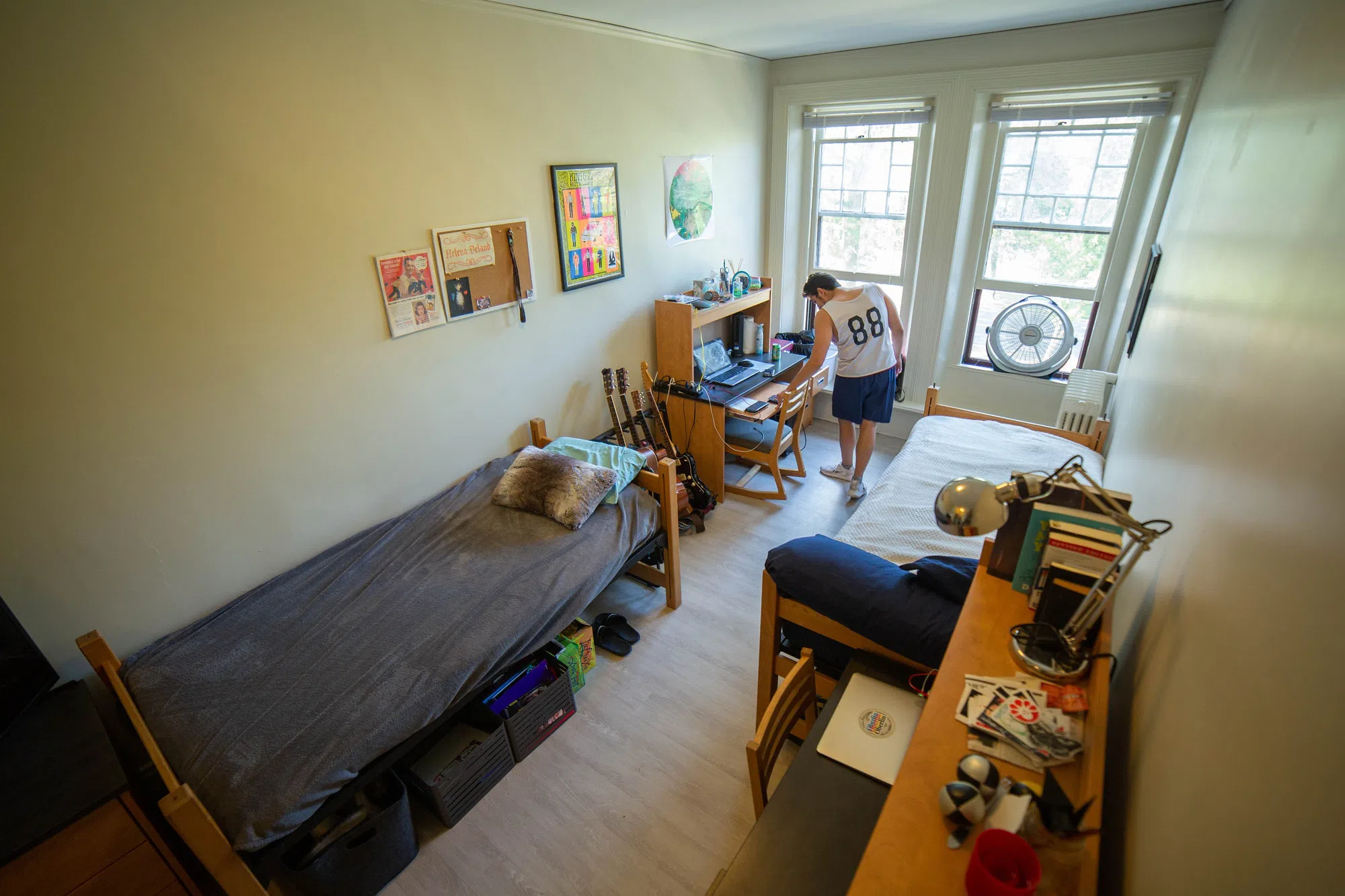 A student moves a chair in his dorm room, filled w 2 beds, 2 desks, belongings, and light streaming in from the 2 large windows. 