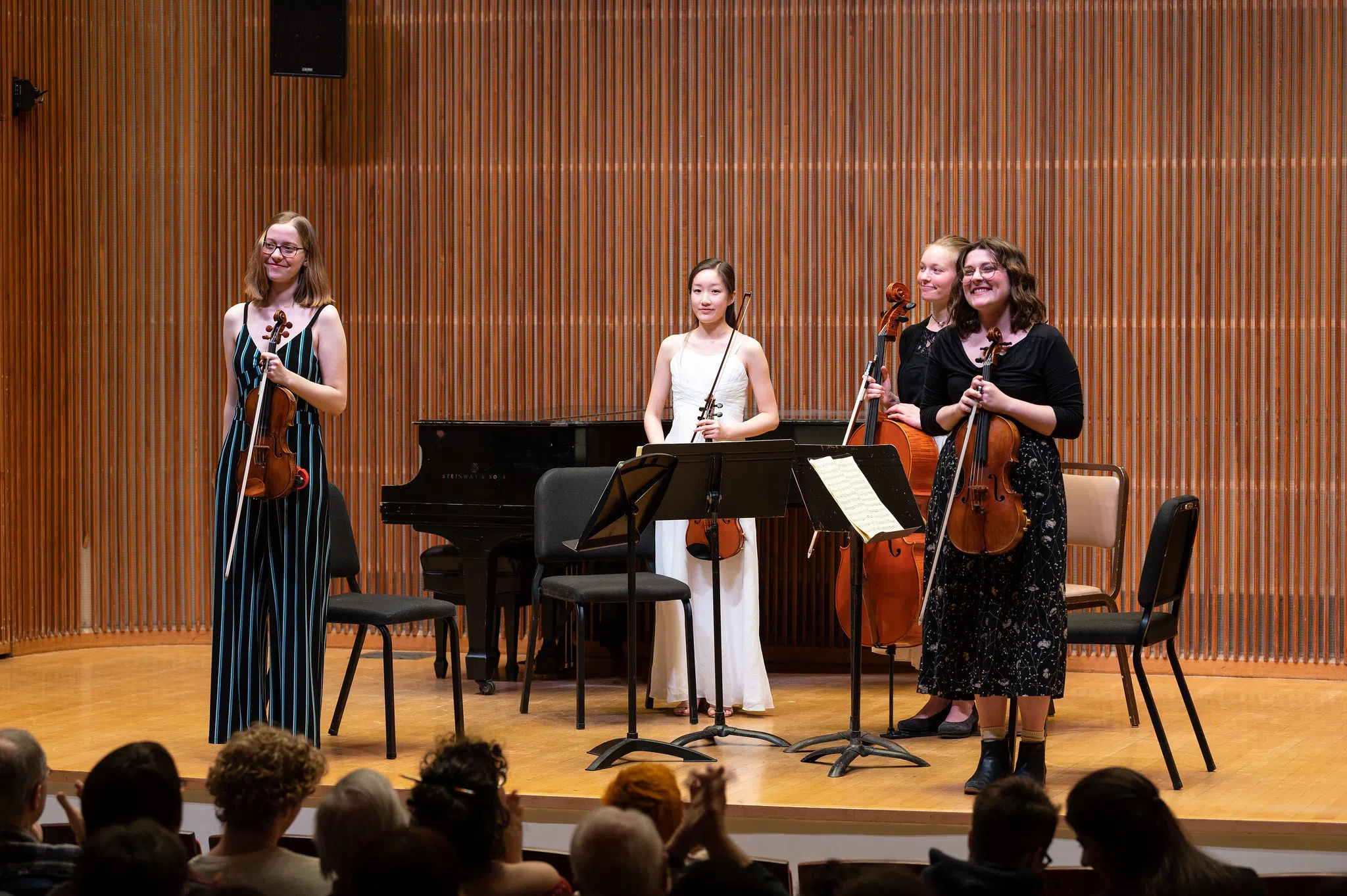 Four students holding stringed instruments stand before an audience on a stage.
