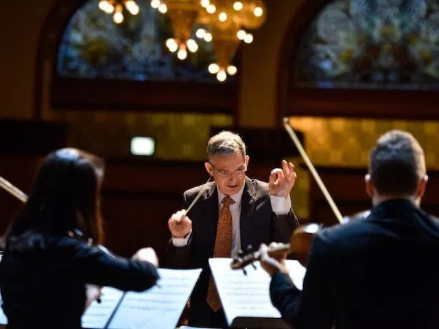 A conductor holding a baton elegantly conducts a group of orchestra students in Finney Chapel.