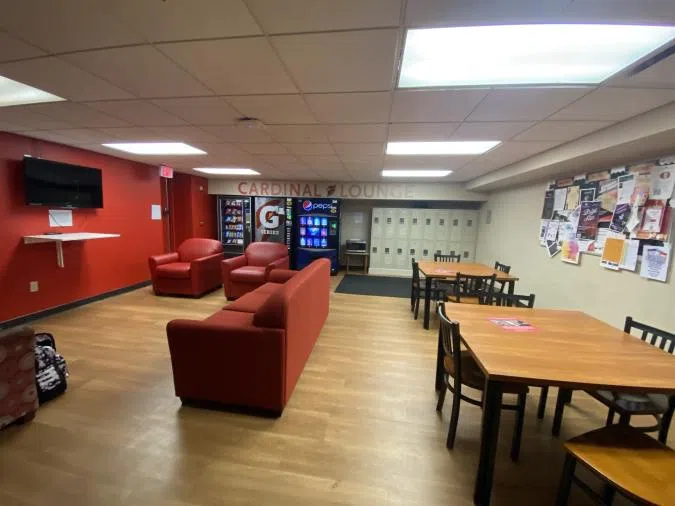 A room with beige walls, and a red accent wall. There are lockers in the right corner, and vending machines in the left. There are couches and tables with a TV on the red wall.