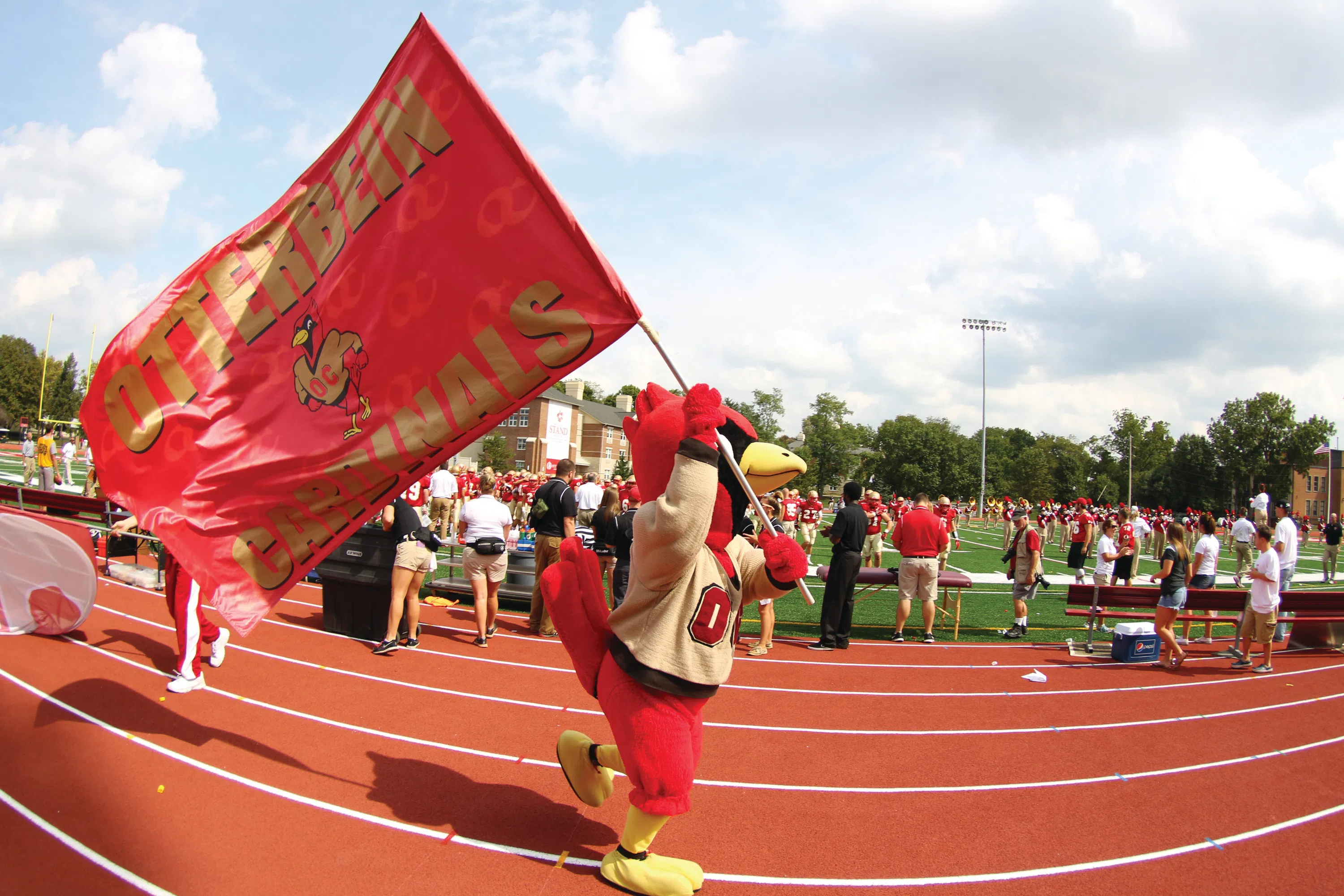Cardy, Otterbein's Athletics Mascot runs on the track carrying our Athletics flag as a crowd looks on.