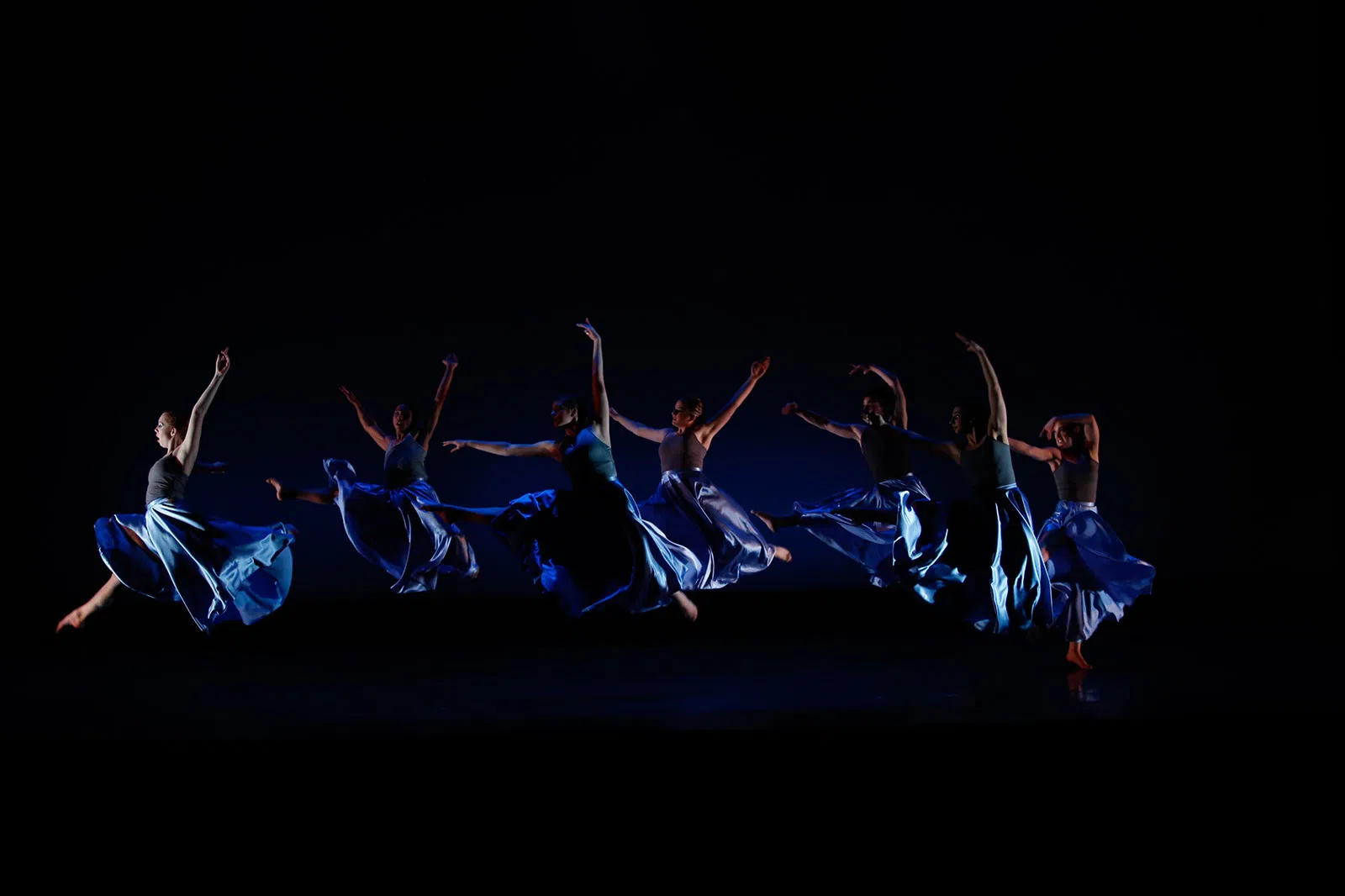 7 dancers are preforming. They are dressed in blue under low lighting.