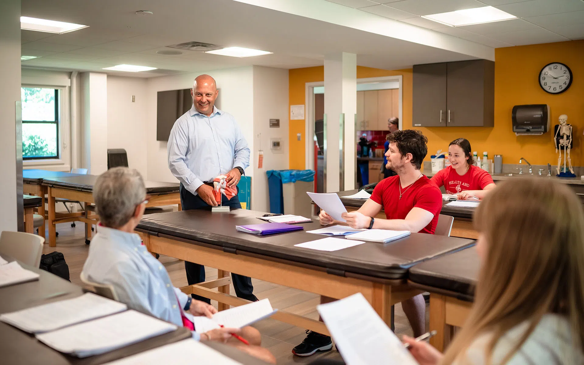 Classroom of students in a lab setting. There is a professor assisting students.