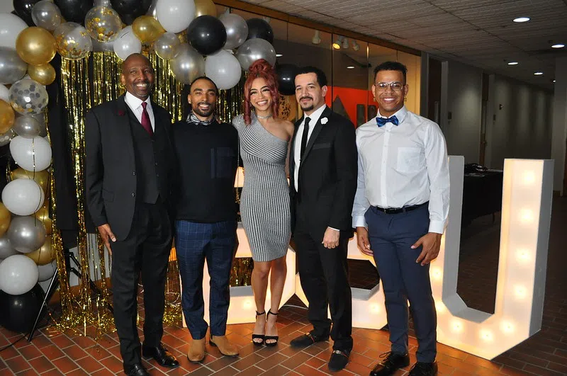 A group of people in formal wear at a ball smiling for a photo. The backdrop is full black, gold, silver, and white balloons.