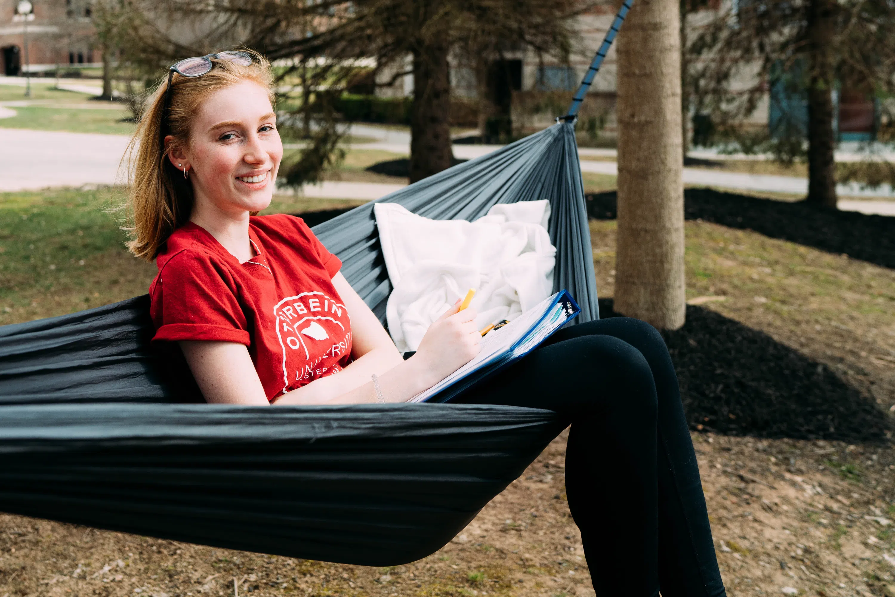 Student sits in a hammock hung in an outside area near a residence hall and reads.