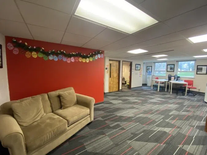 A lobby area with a red accent wall and beige walls. There is a tan couch directly infront, with a desk in the back of the room. There is a large copier and printer to the right o the desk.