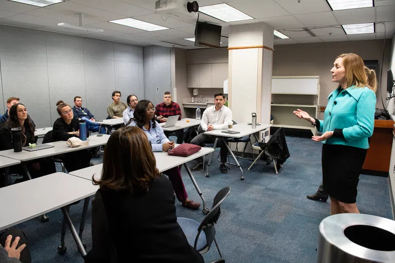 Female instructor stands in front of a class of students. She is speaking and gesturing with her hands. 