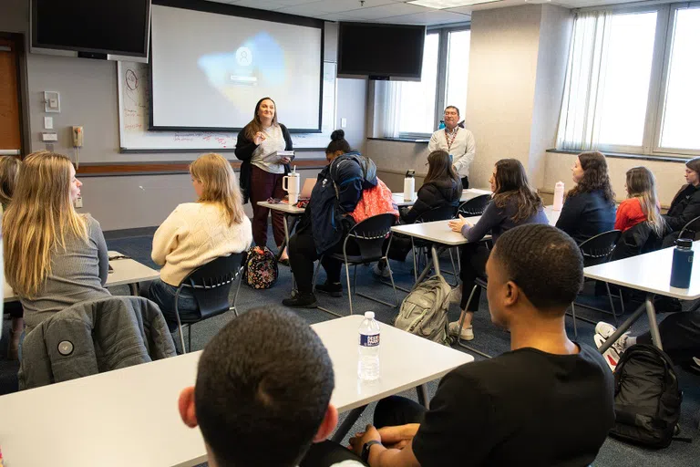 A woman stands at the front of a classroom full of students sitting at tables. Instructor stands off to the side of the room. 