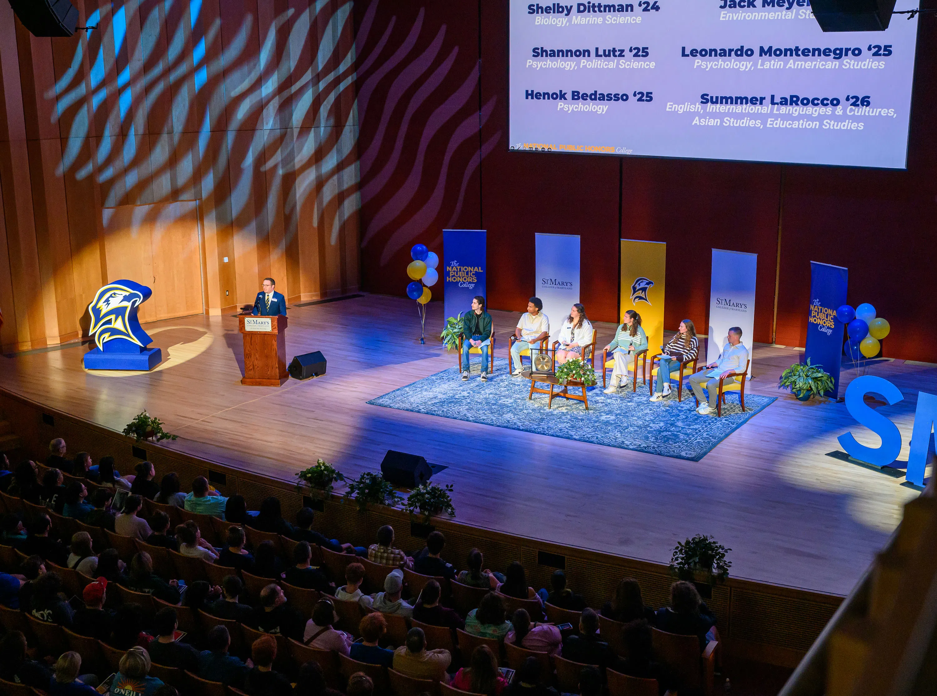 Concert hall stage decorated for Admission's event. All visible chairs in the audience are filled and six students are seated around a coffee table for a panel moderated by the Director of Admission on stage.