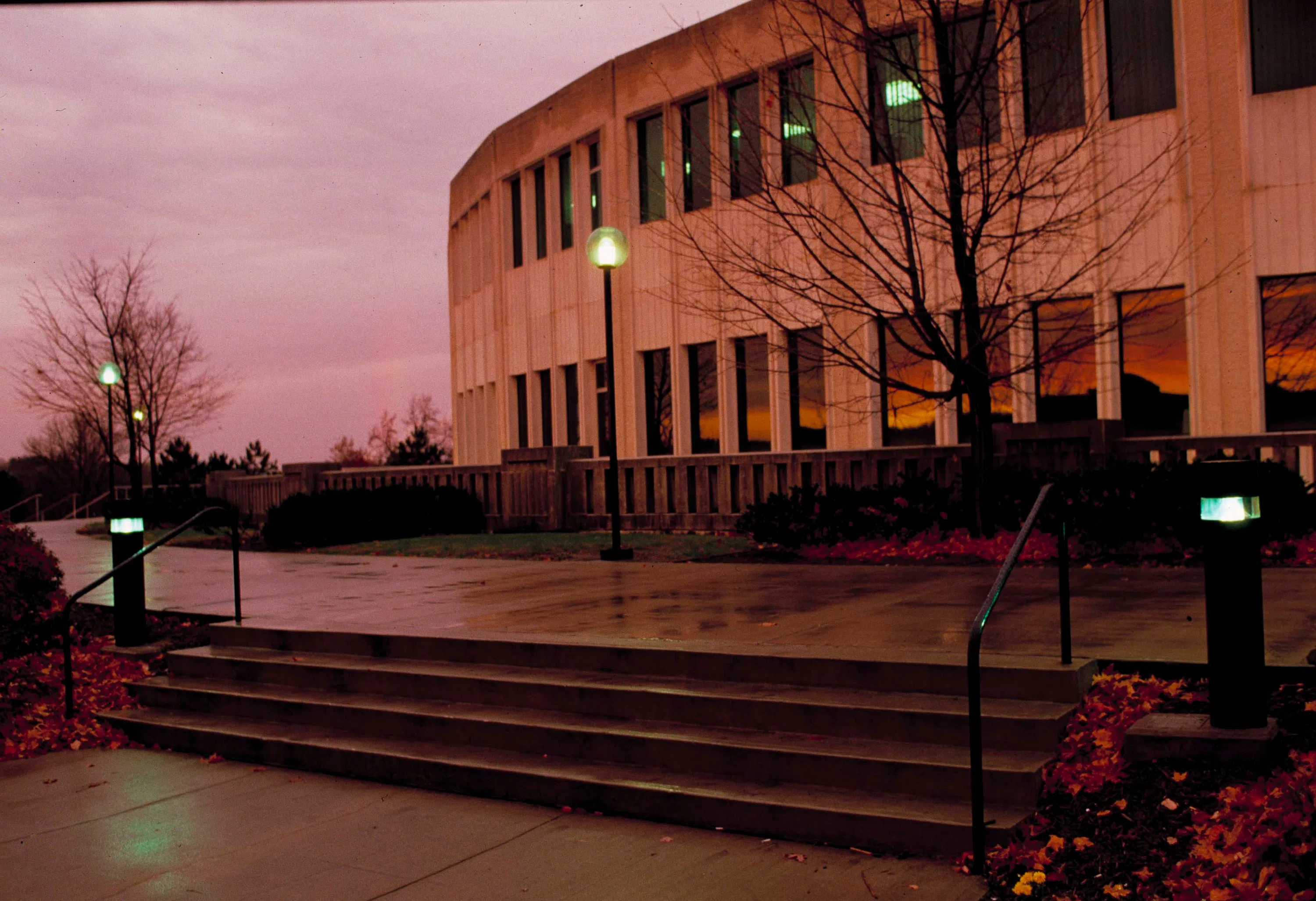 A curved building with small, dark windows is featured in front of a light pink sunset.