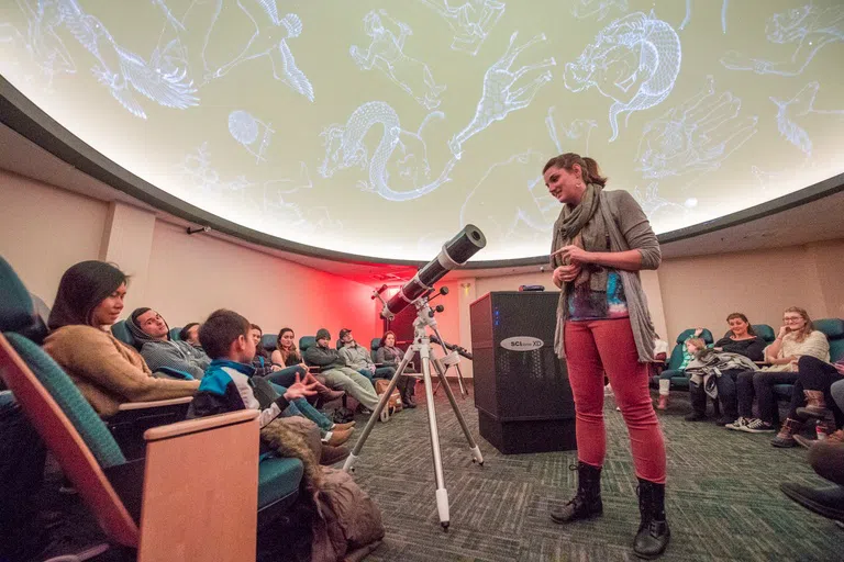 Students are seated in the planetarium gazing up at the screen while an instructor points out the constellations.