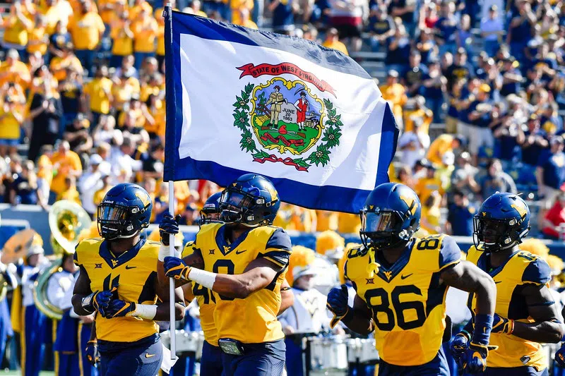 WVU football players charge the field carrying the flag of the state of West Virginia.