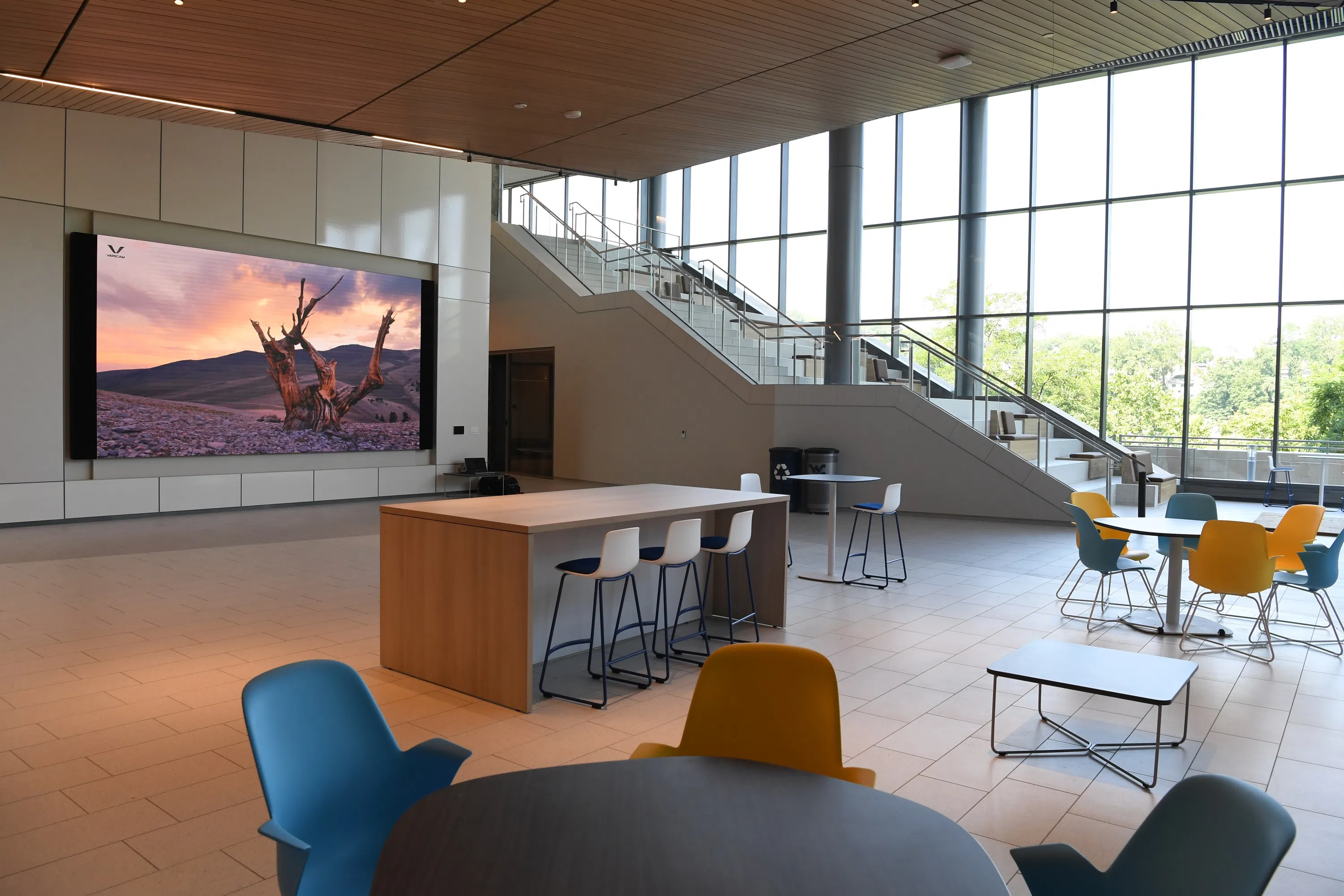Image shows open room with various desk and chair setups for students to use. There is a large screen display next to the grand staircase, complete with stair seating. 