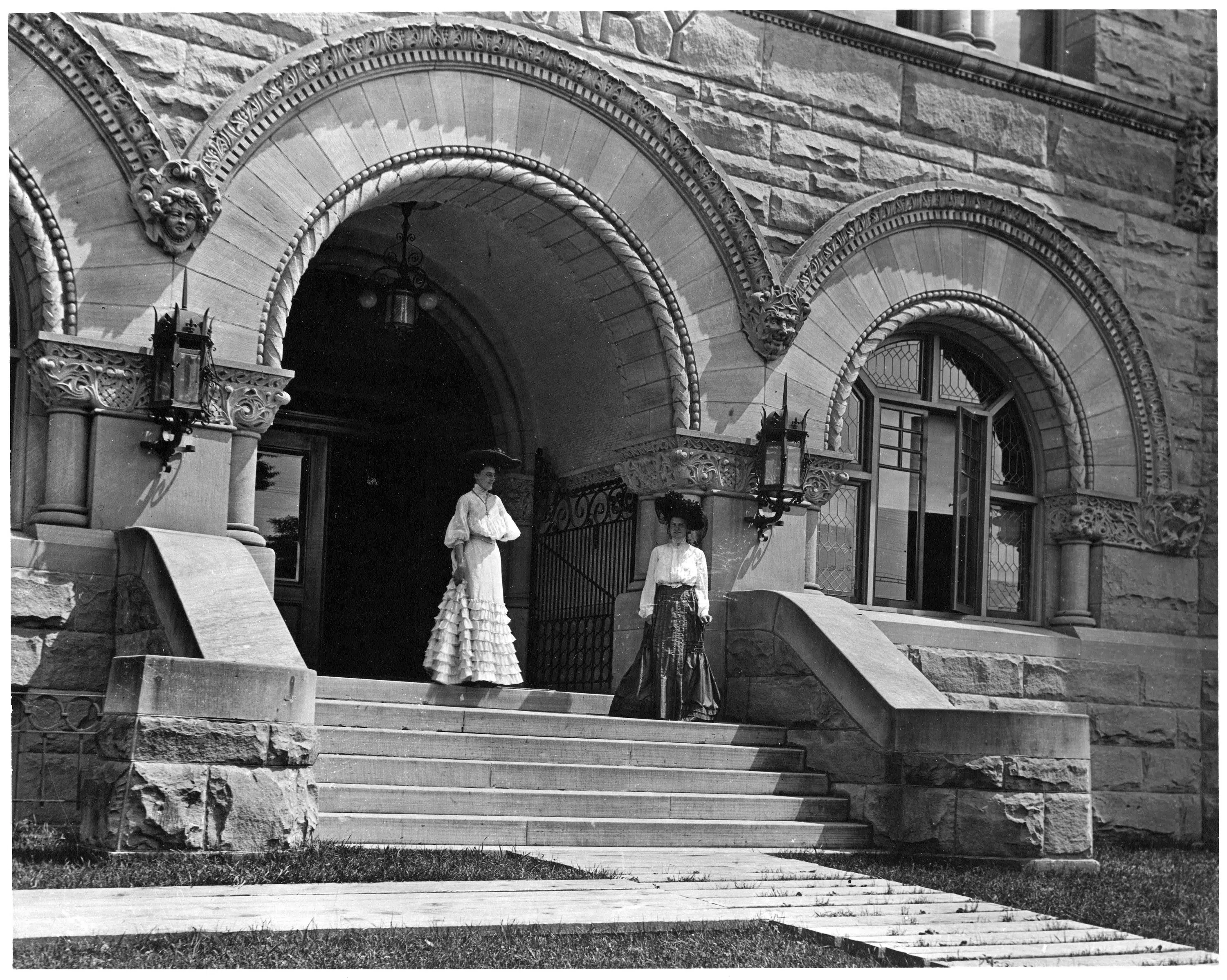 A black and white photograph shows two women stand on the steps of Stewart Hall c. 1905