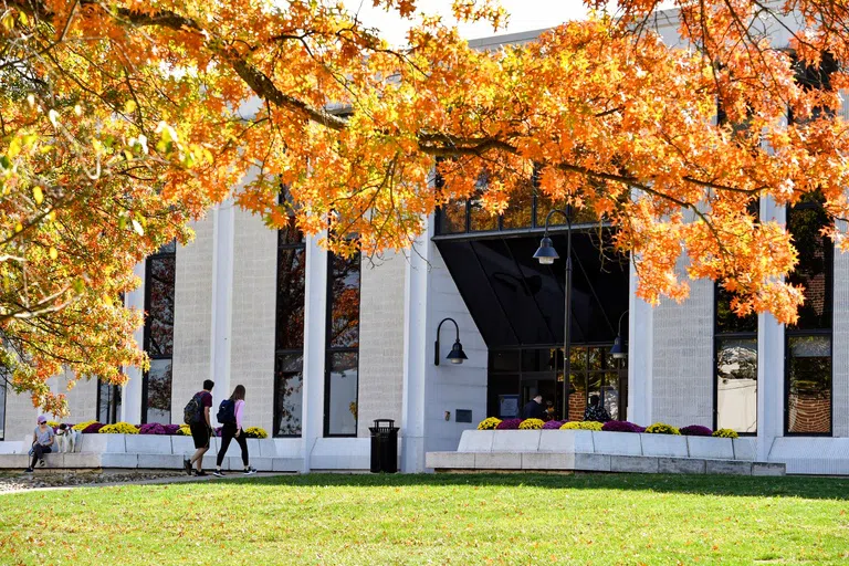 Image shows the exterior of the Evansdale Library, a light brick building with tall, black windows and students filing in and out. 