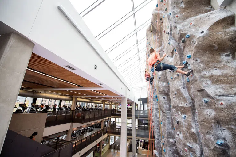 Student climbing indoor rock wall with various cardio equipment visible in the background