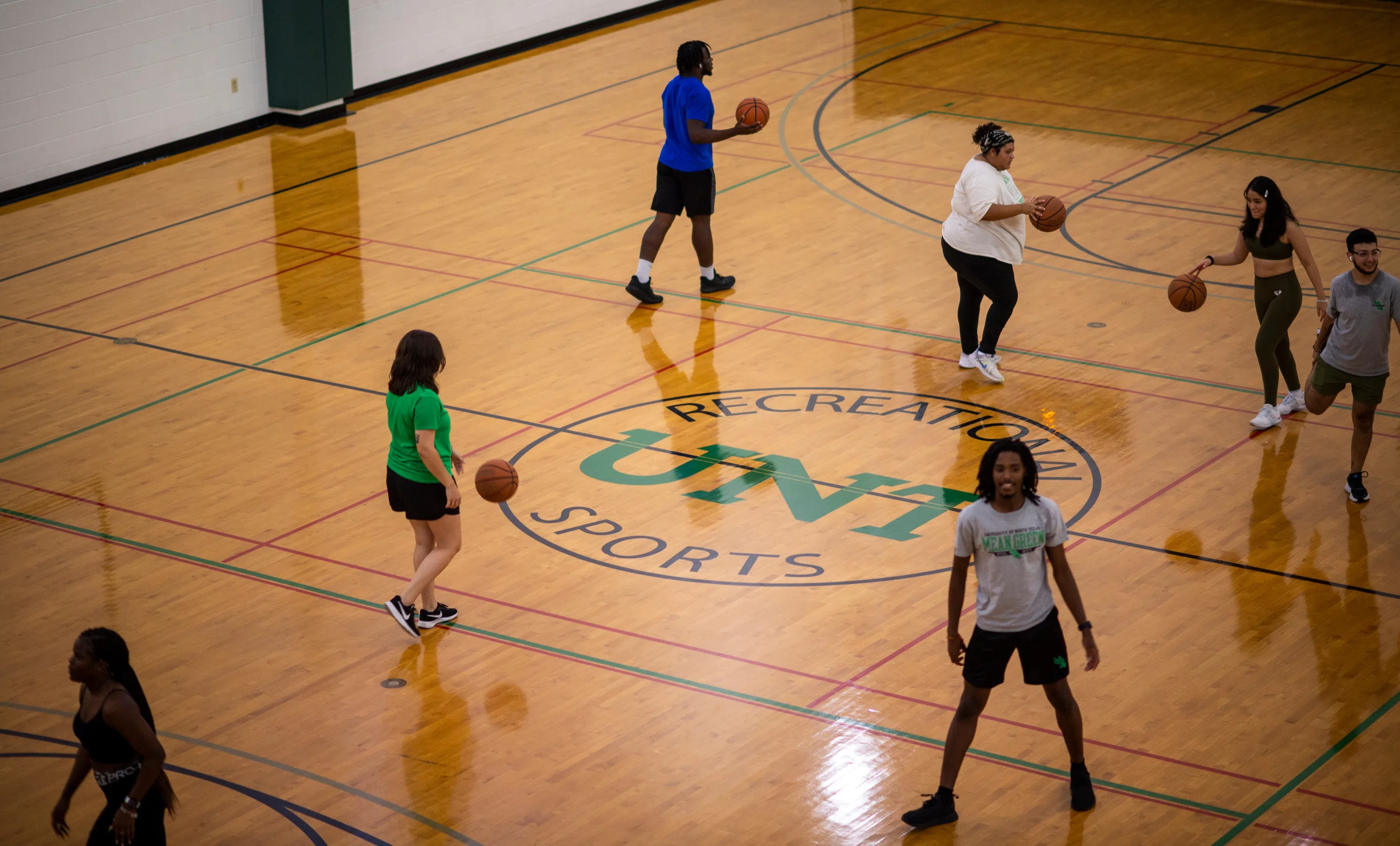 Students play with their own basketballs on a basketball court. 'UNT' is printed on the center of the court's floor. 