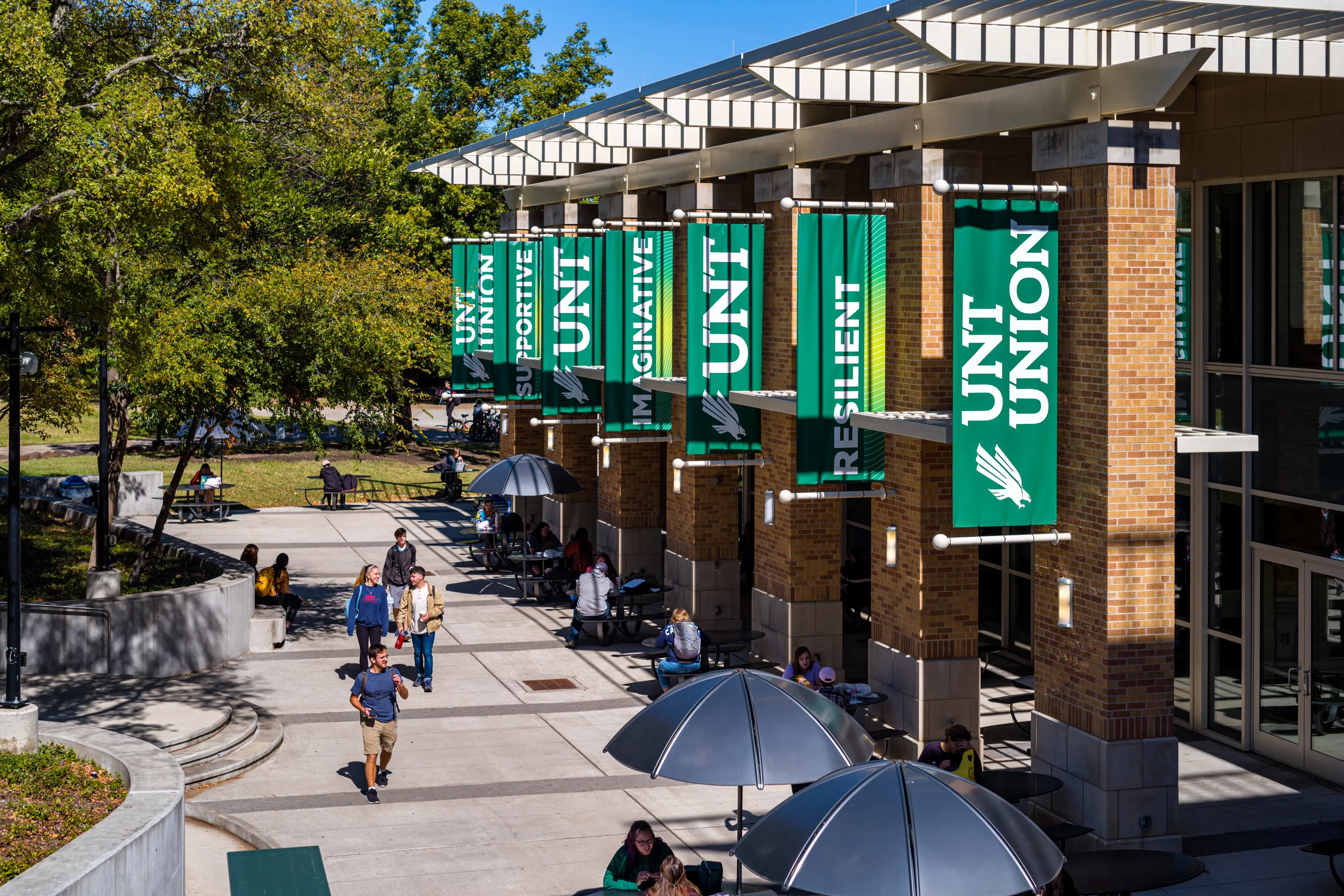 Exterior of University Union. UNT banners hang from the side of the building, students study under shaded outdoor tables, and people walk on the sidewalk. 