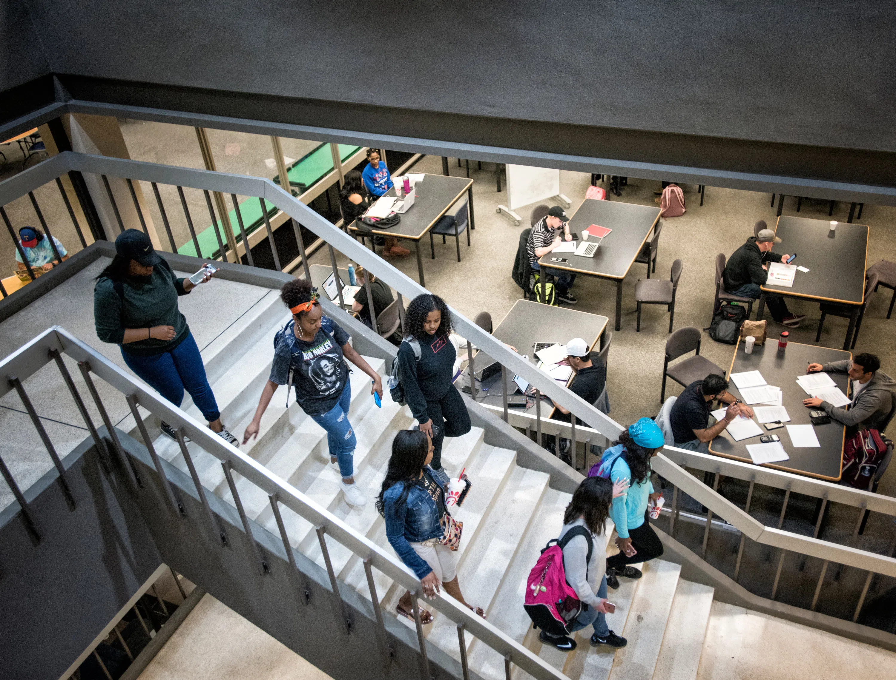 A group of students walk down a staircase in Willis Library. Students studying at communal tables can be seen in the background. 