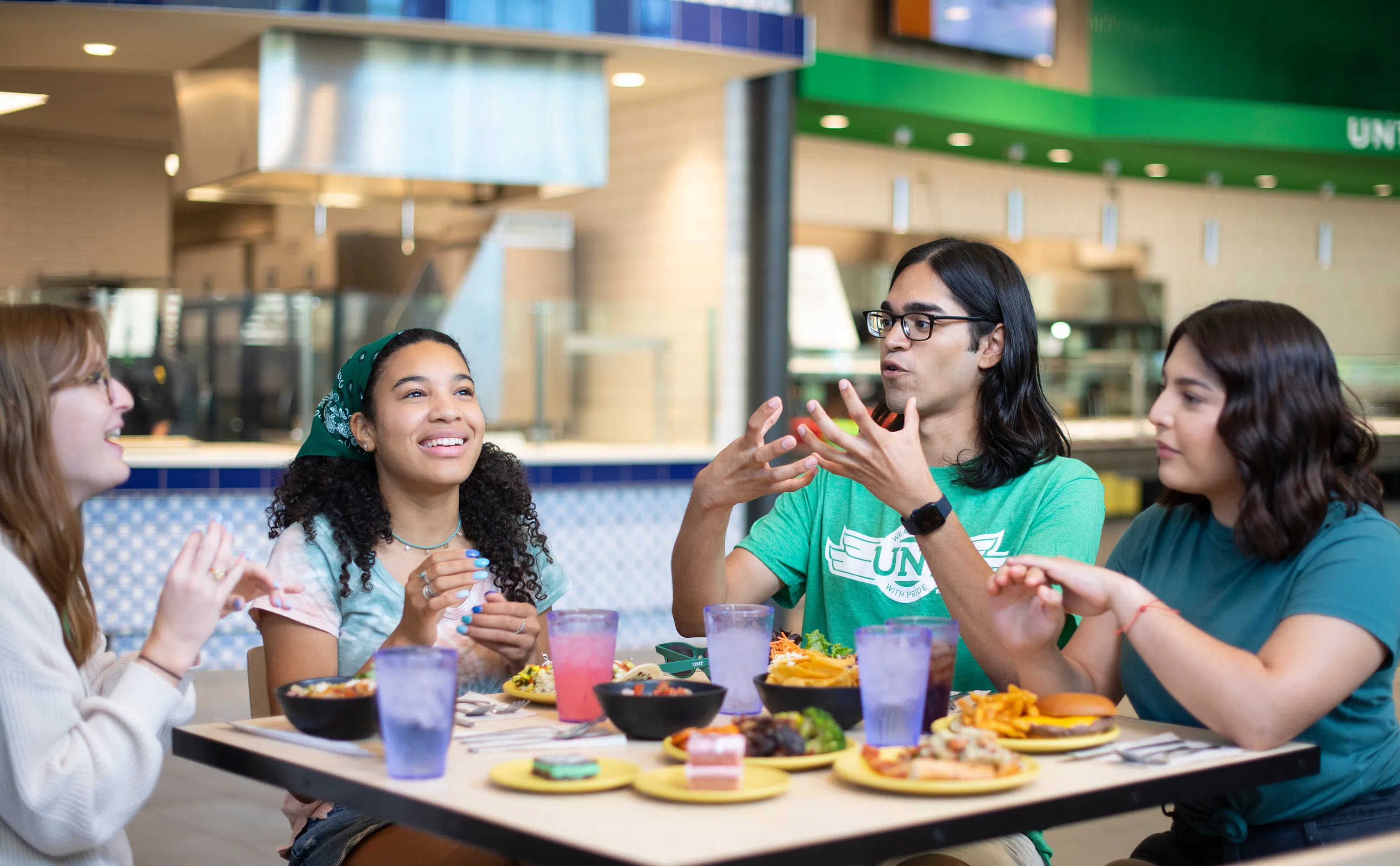Four students chat and smile enthusiastically while sitting at a table with various food choices from the different places at Eagle Landing. 