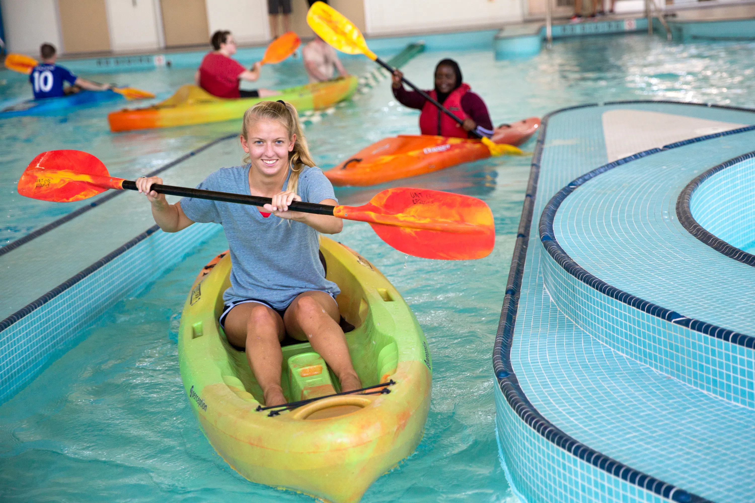 Student in a kayak in a pool. The student is paddling the kayak and smiling at the camera. 
