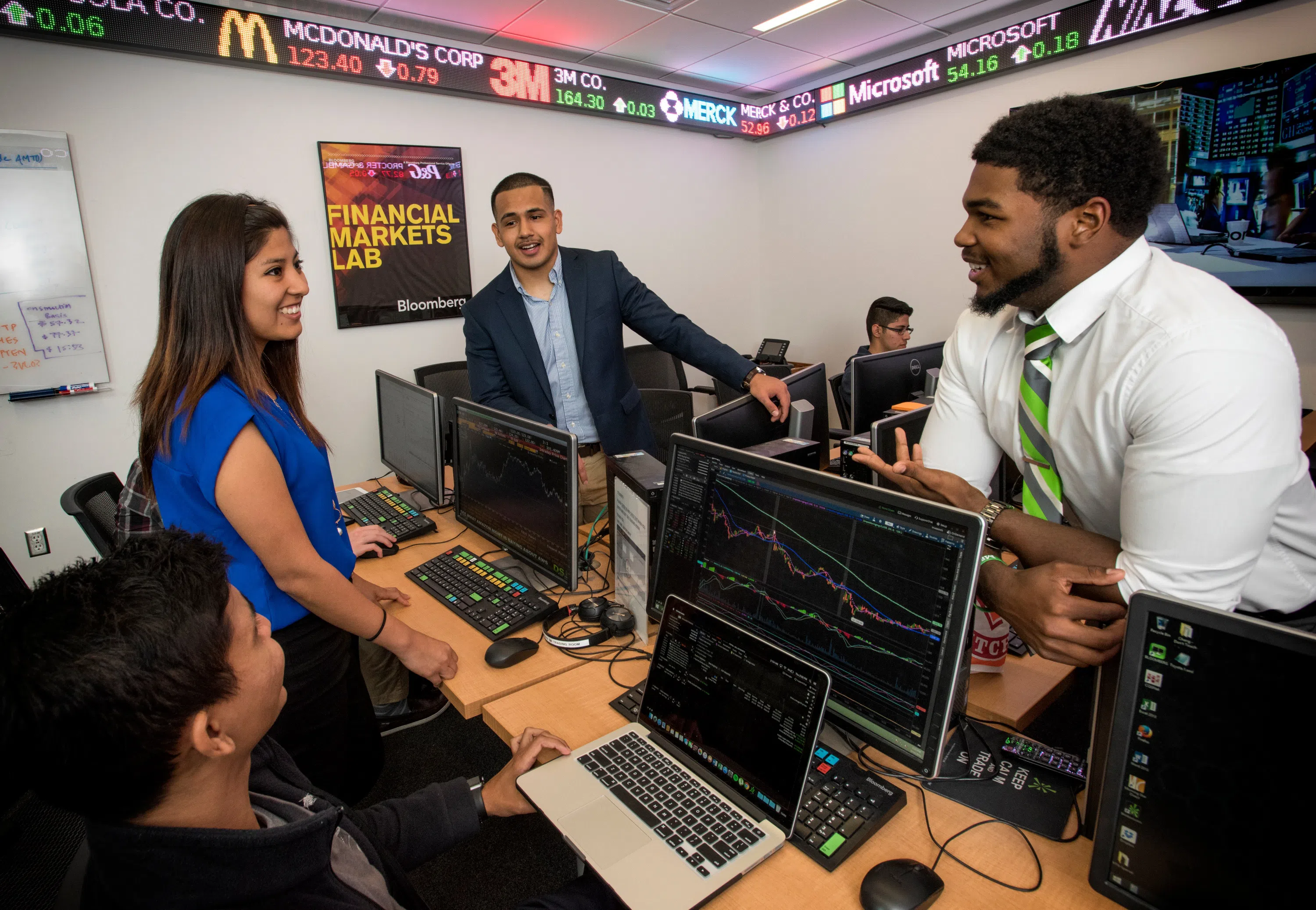 Students gather around computers displaying stock information. Stock ticker runs around the top of the room, displaying different stocks' performance