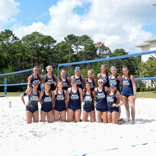 women's volleyball team pose on sand court in two rows, one row kneeling in front of the other