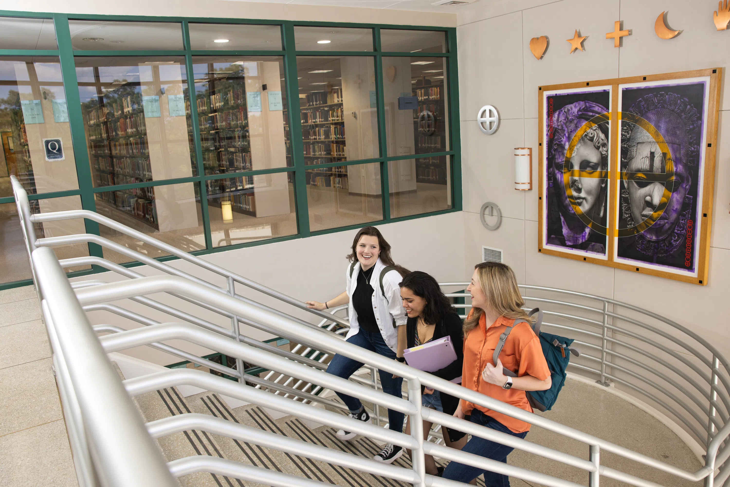 Three female students walking up stairs in the library with large art pieces hanging in the background.