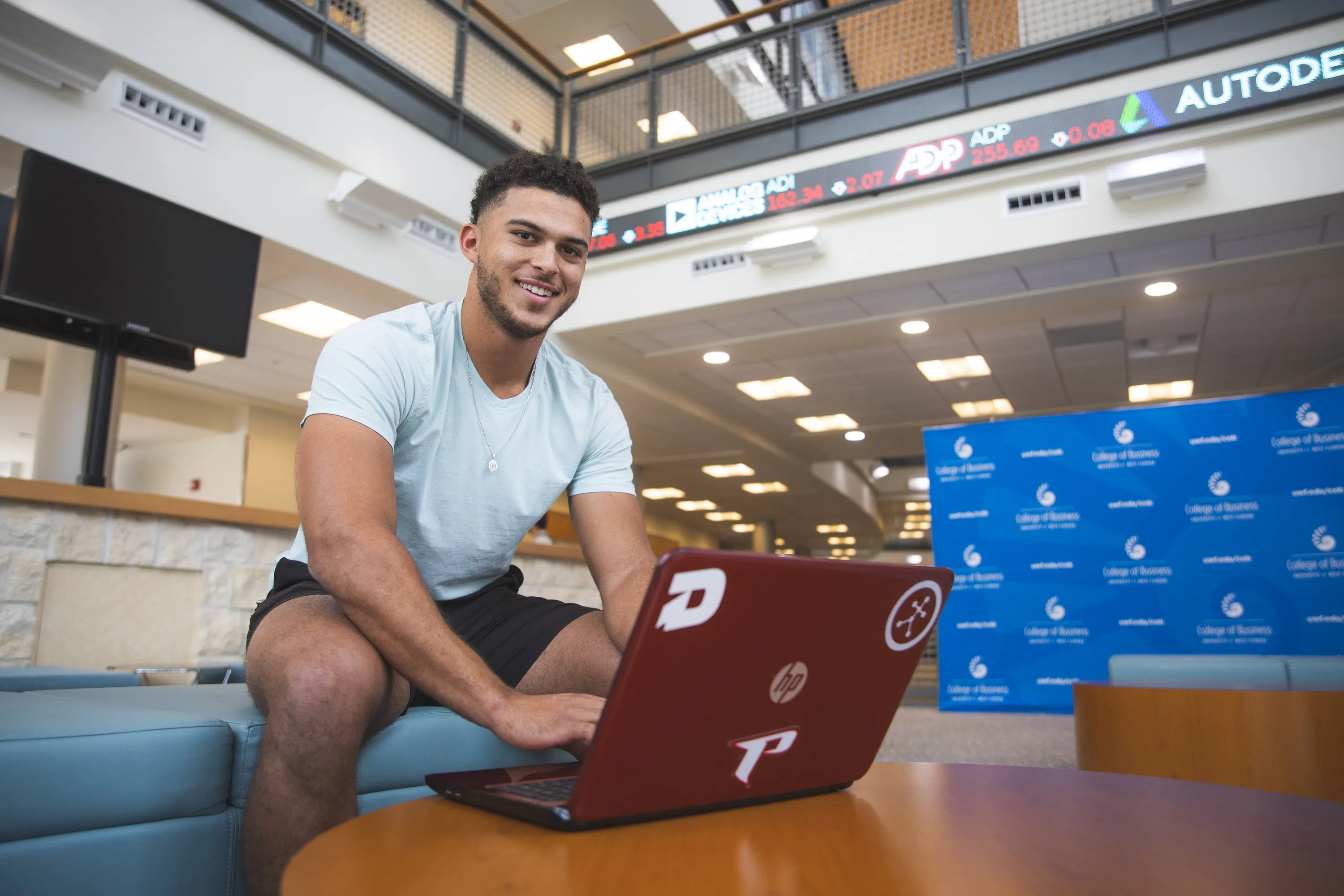 Student on laptop in the Lewis Bear Jr. College of Business Atrium with the New York Stock Exchange Ticker in the background. 