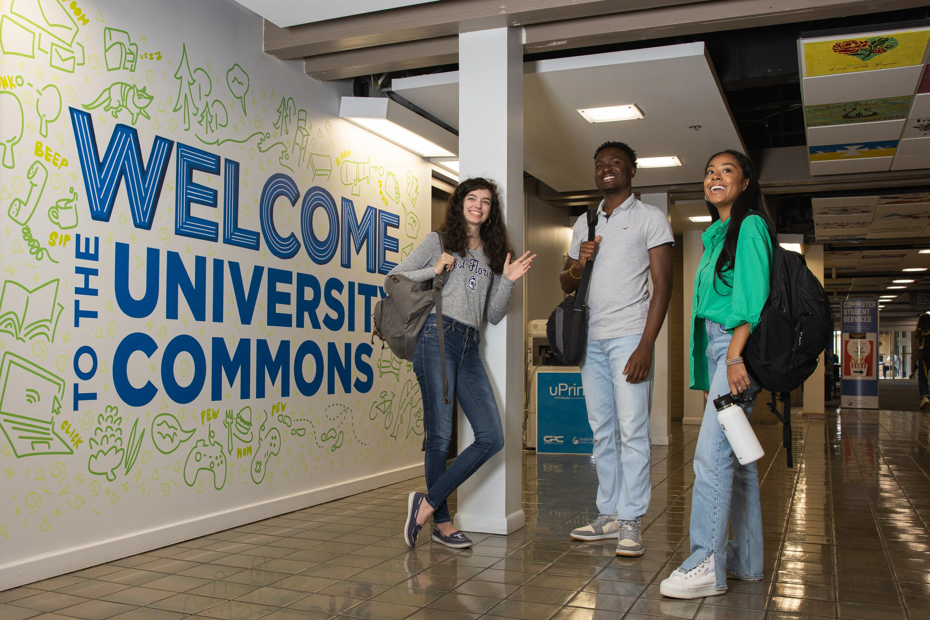 Three students standing in front of Welcome to the University Commons mural outside of the Nautilus Market
