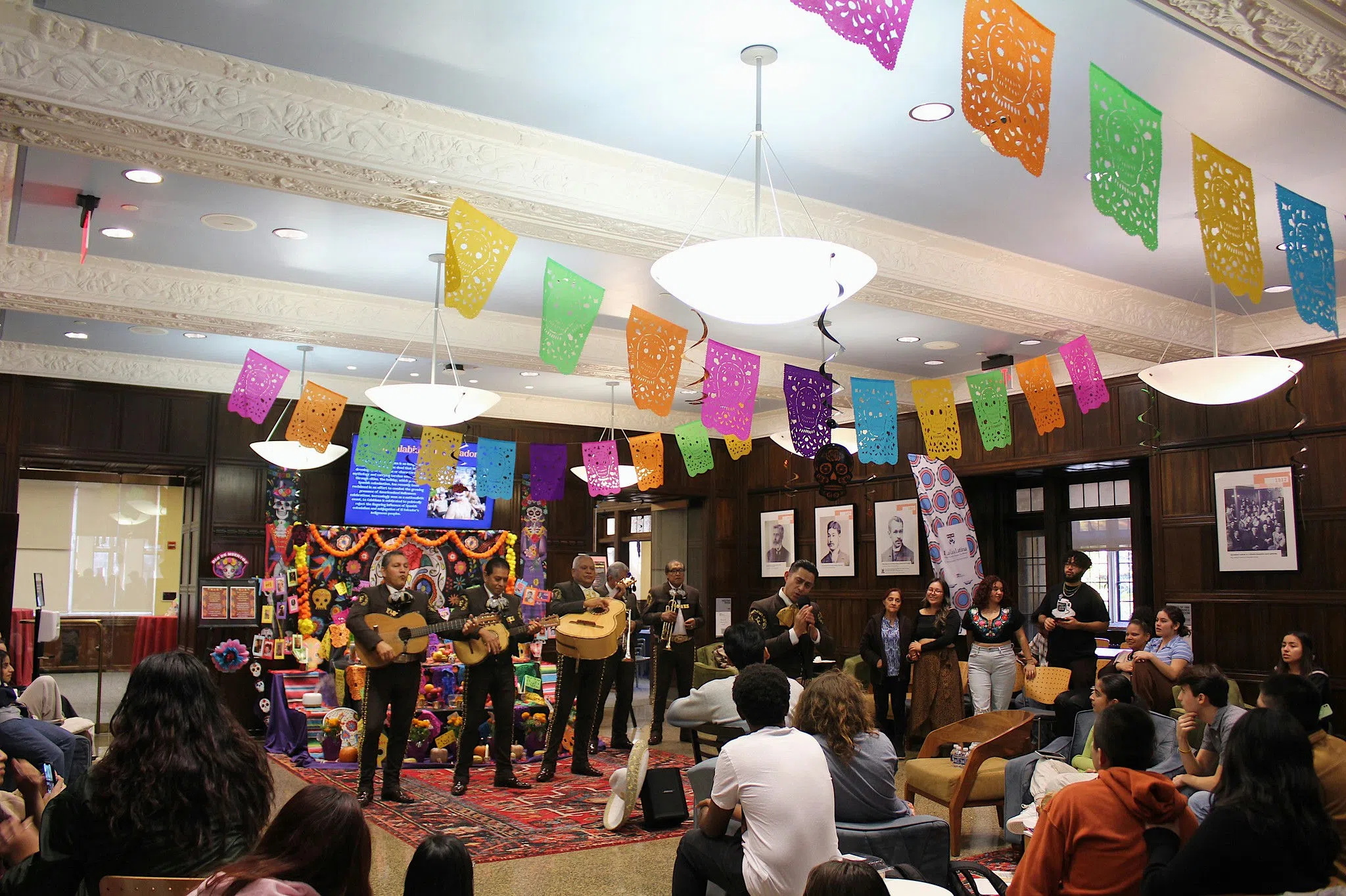 A group of students are listening to a Mariachi band in traditional clothing. They are gathered in a large space decorated with colorful paper garlands where a large ofrenda (or altar) has been constructed, which is customary for the Day of the Day. 